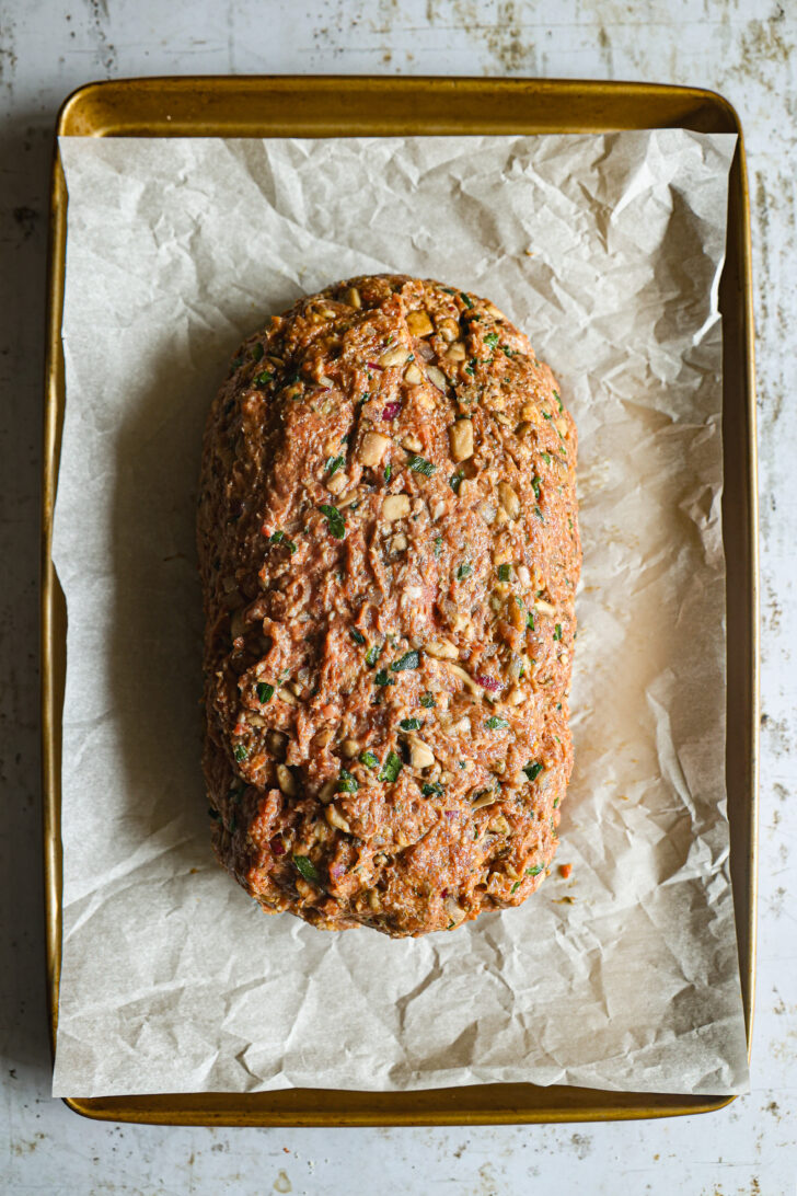 A raw meatloaf packed with diced vegetables and herbs, shaped into a loaf and placed on a parchment-lined baking sheet.