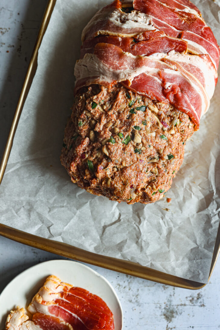 A partially wrapped meatloaf with bacon strips layered on top, placed on a parchment-lined baking sheet with extra bacon on a plate nearby.