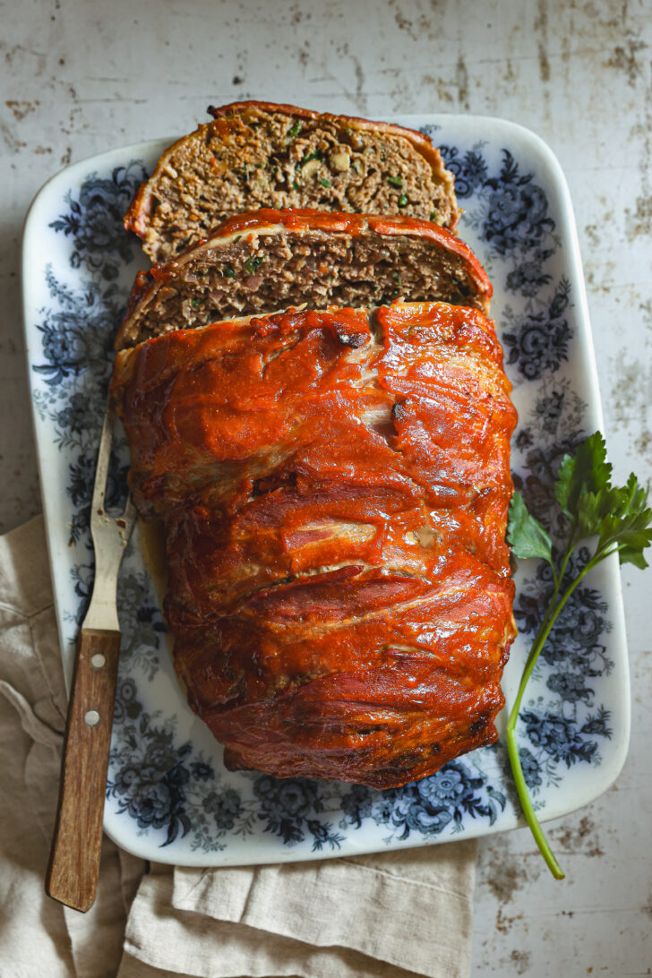 A beautifully baked meatloaf wrapped in bacon, sliced to reveal its tender interior, served on a floral platter with parsley garnish.