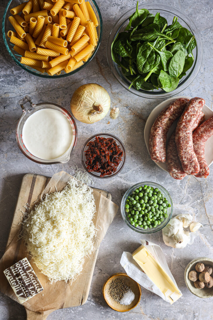 Ingredients for Creamy Tuscan Rigatoni, including pasta, spinach, cheese, peas, sausage, and spices, arranged on a countertop