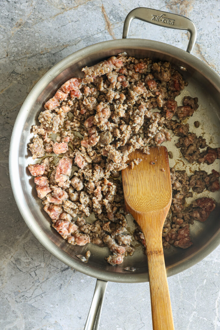 Browned Italian sausage cooking in a stainless steel skillet with a wooden spatula resting on the edge of the pan.