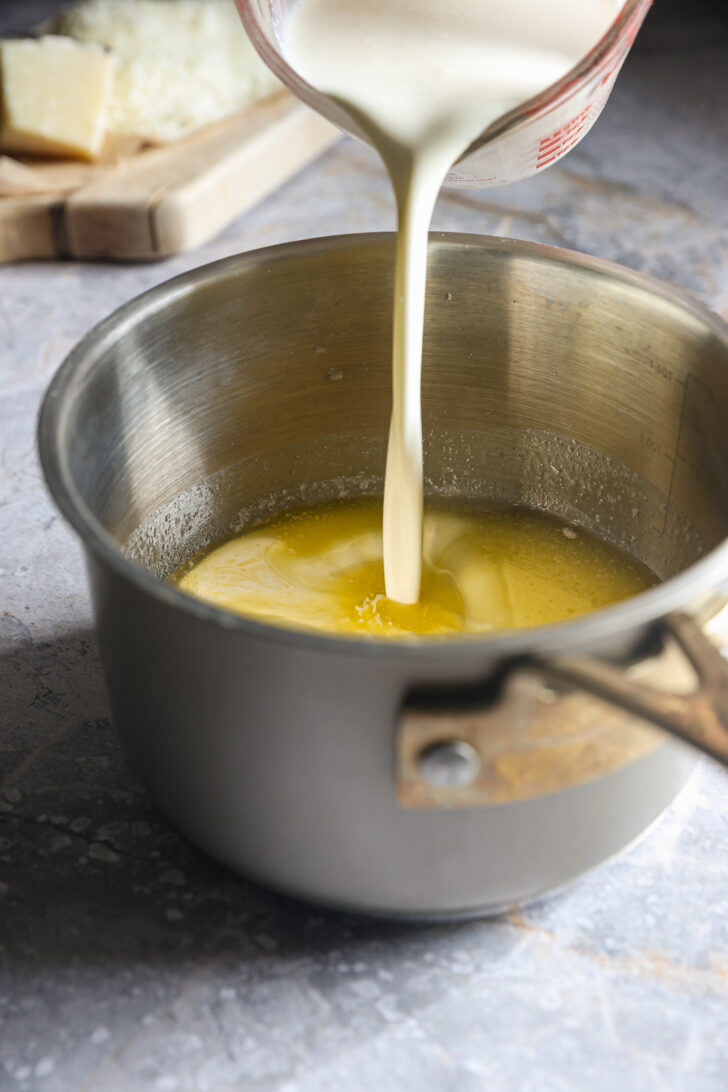 Heavy cream being poured into a saucepan of melted butter, preparing a rich Alfredo sauce for the dish.