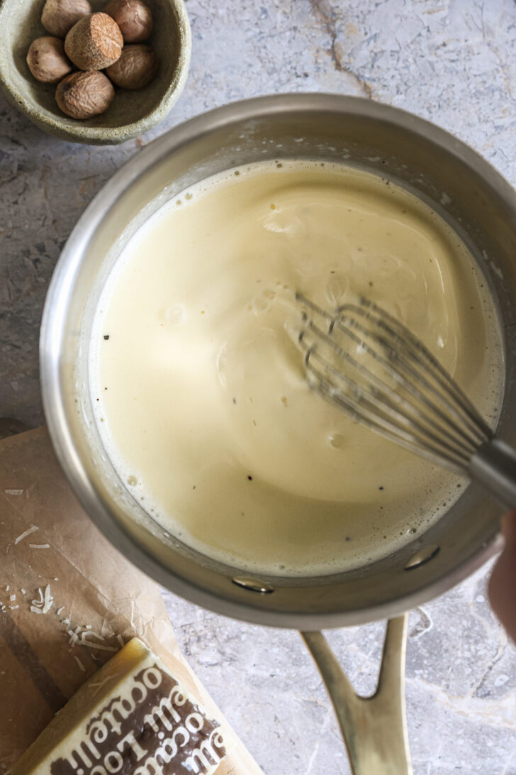 Rich Alfredo sauce being whisked in a saucepan until creamy and smooth, with grated cheese and whole nutmeg nearby for flavor.