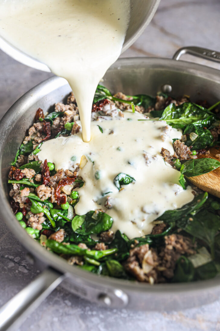 Creamy Alfredo sauce being poured over a skillet of cooked sausage, spinach, sun-dried tomatoes, and peas for a rich pasta dish.