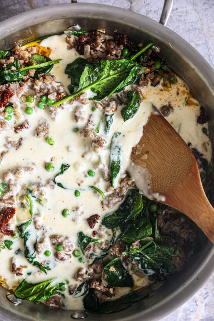 A skillet filled with creamy Alfredo sauce, sautéed spinach, peas, sun-dried tomatoes, and sausage, mixed with a wooden spatula.