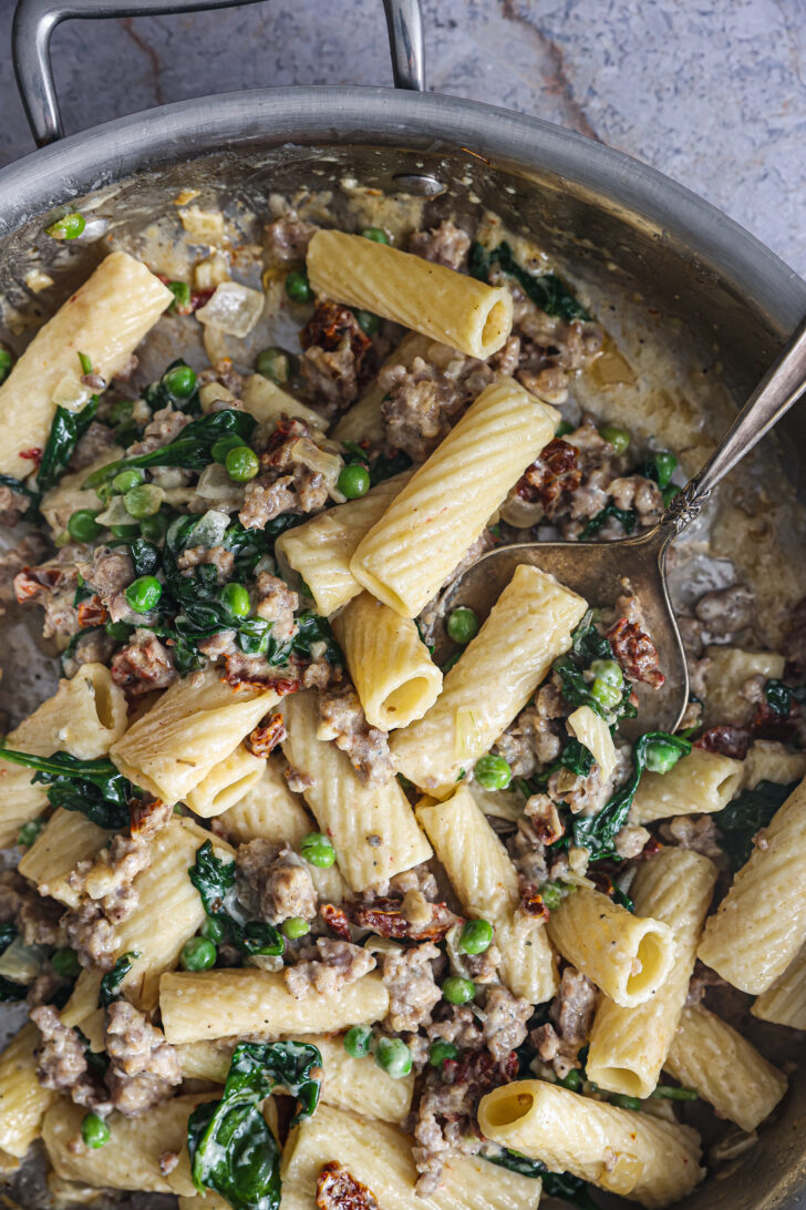 Creamy Tuscan Rigatoni in a skillet, featuring tender pasta, sausage, spinach, peas, and sun-dried tomatoes, ready to serve