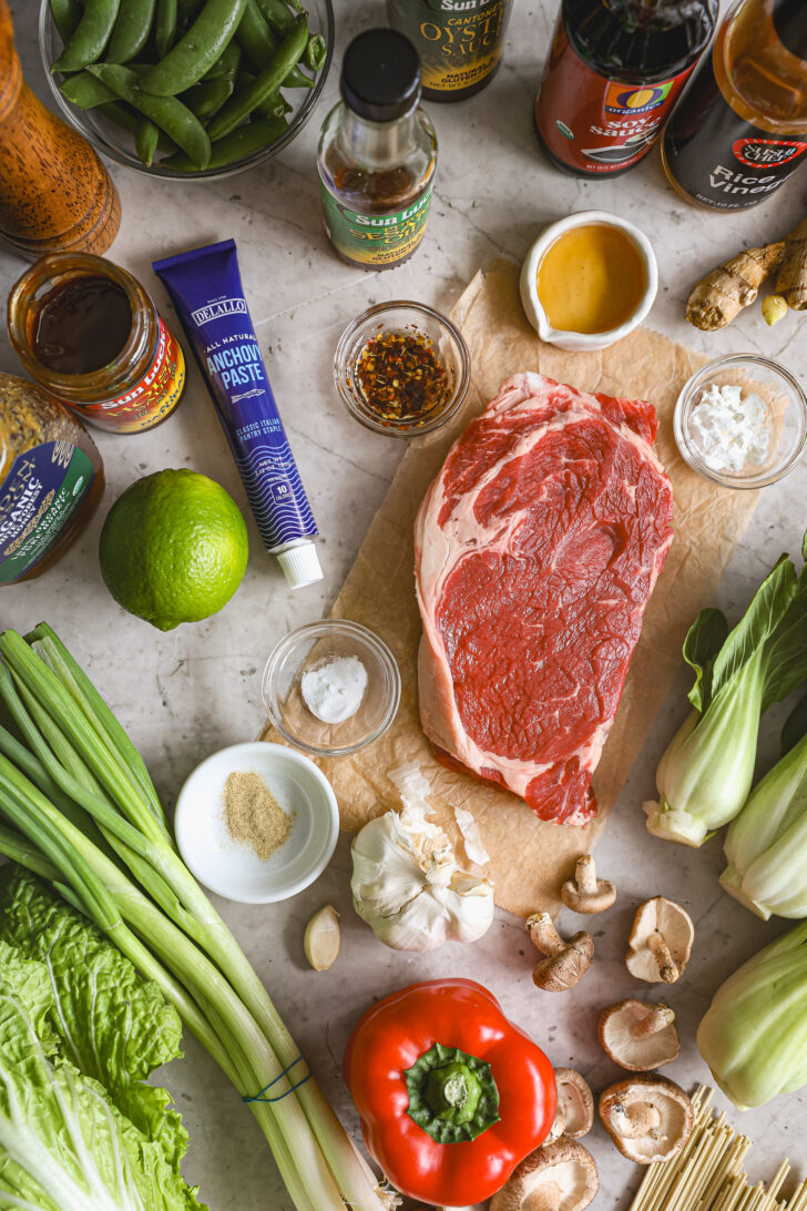 Fresh ingredients for beef lo mein, including ribeye steak, bok choy, red bell pepper, scallions, mushrooms, sauces, and seasonings on a countertop