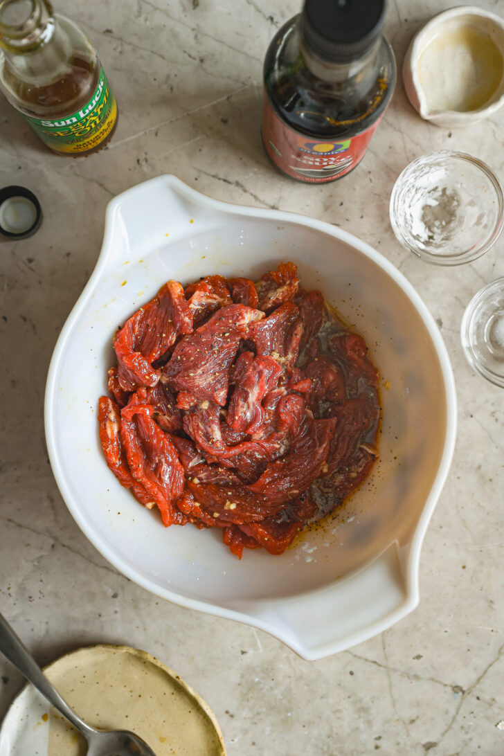 Marinated beef strips in a white bowl with soy sauce and seasonings, surrounded by sauce bottles and empty bowls on a marble countertop.