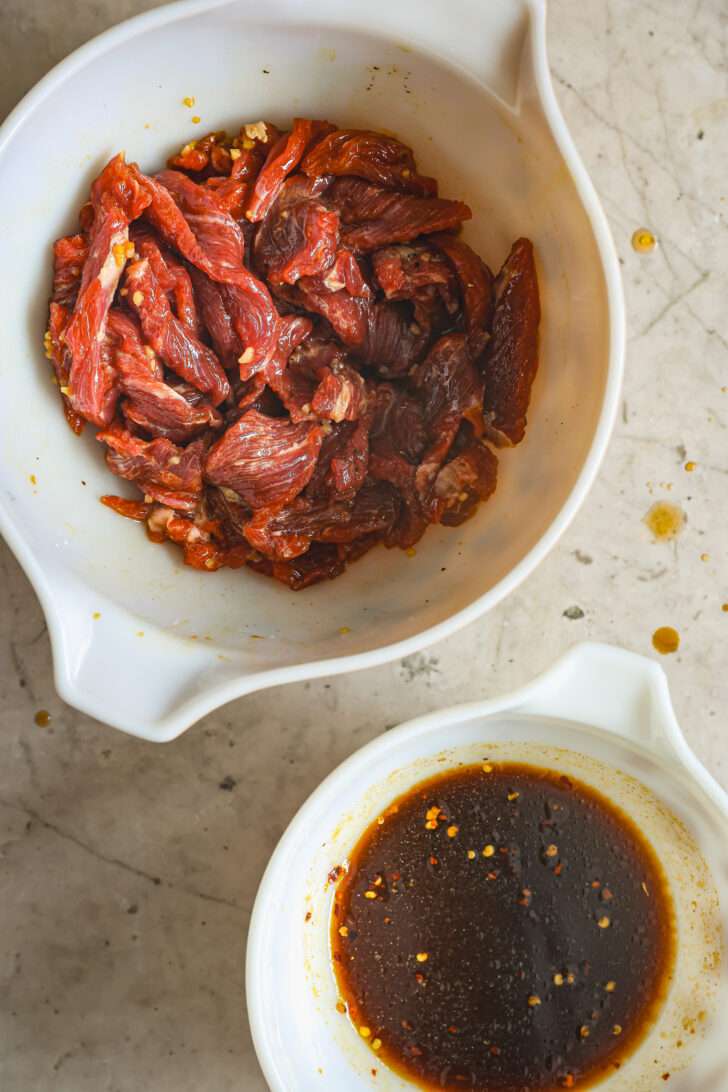 Marinated beef in a white bowl with a separate bowl of dark soy-based sauce with chili flakes, set on a marble countertop