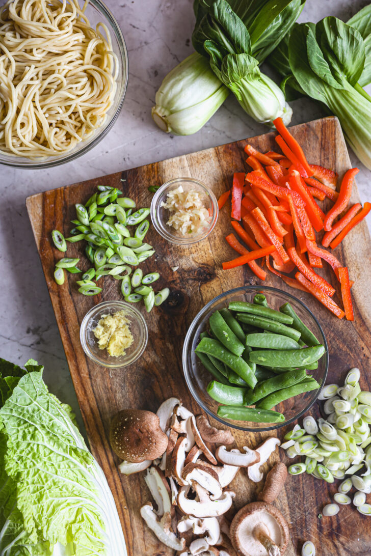 Chopped vegetables, ginger, garlic, and cooked noodles arranged on a wooden board, with bok choy and Napa cabbage nearby