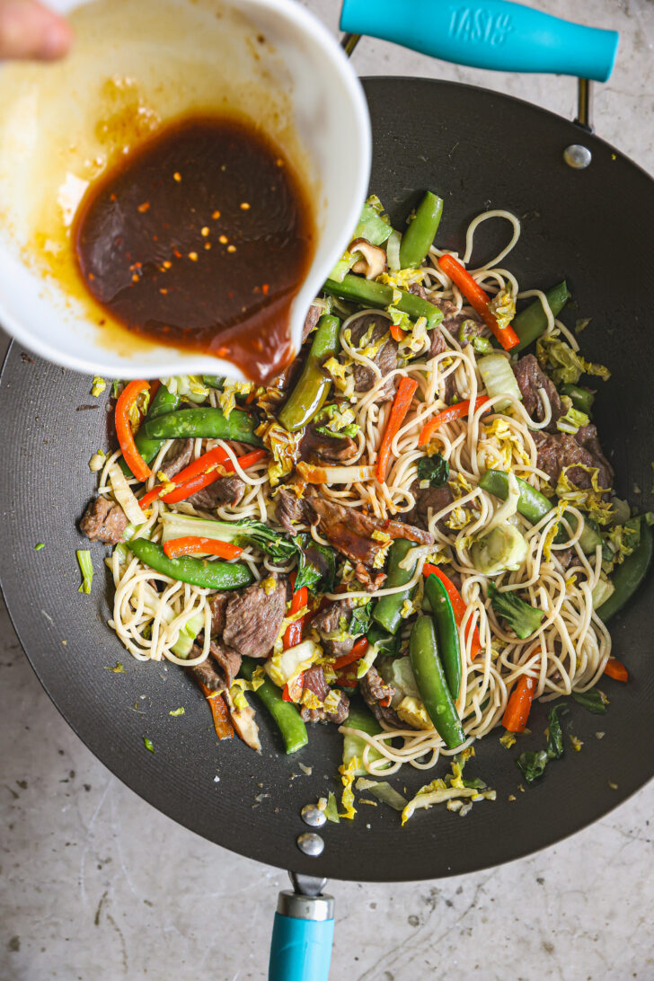 A rich, soy-based sauce with chili flakes is poured over stir-fried beef, snap peas, bell peppers, bok choy, and noodles in a hot wok.