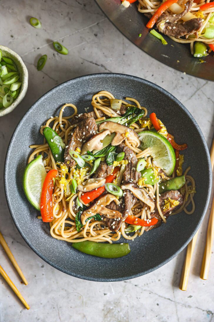 A bowl of beef lo mein with shiitake mushrooms, snap peas, and red bell peppers, garnished with sesame seeds, scallions, and lime wedges