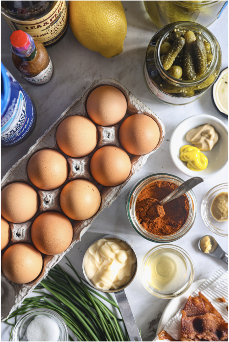 Eggs, paprika, mayo, mustards, vinegar, Worcestershire, chives, bacon, and pickles arranged on a marble surface for deviled eggs.