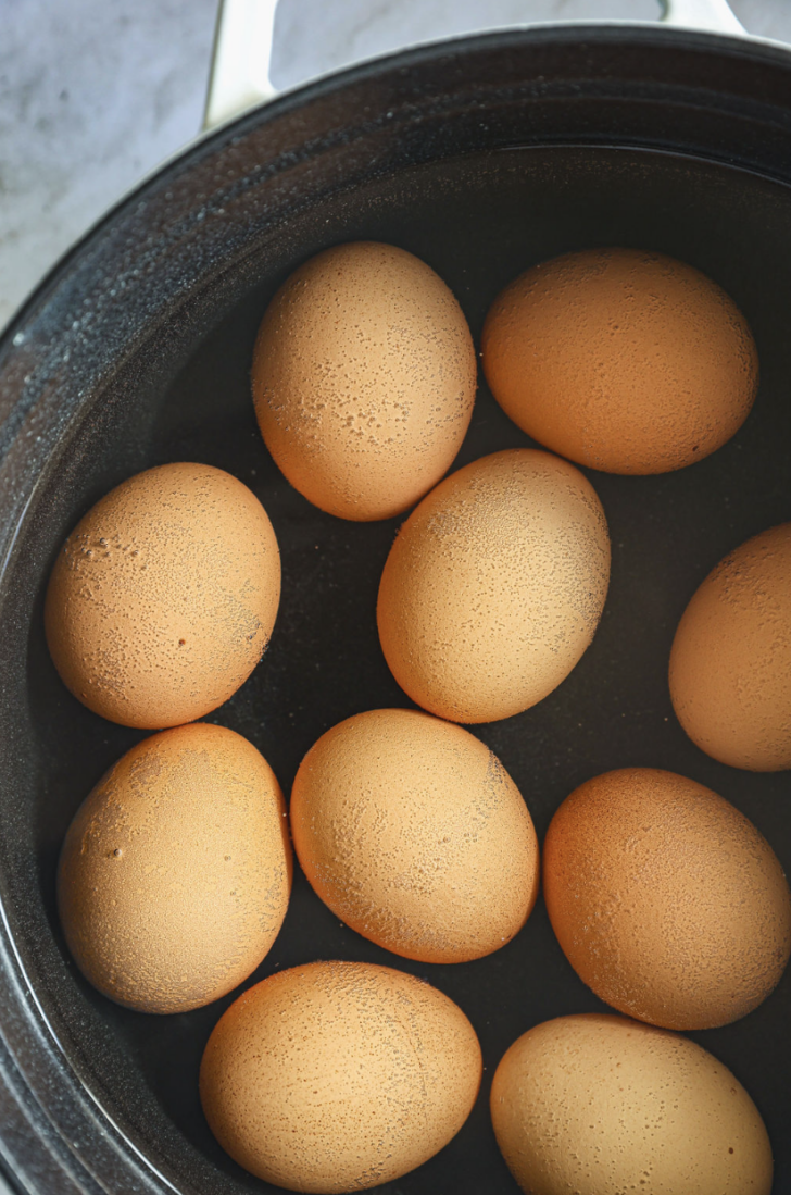 Brown eggs submerged in hot water inside a black pot, beginning the hard-boiling process for deviled eggs.