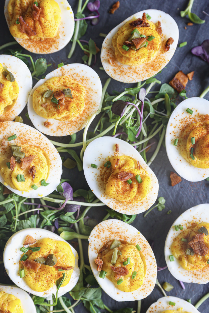 Deviled eggs garnished with paprika, chives, crispy bacon, and pickles, served on a bed of fresh microgreens for extra color