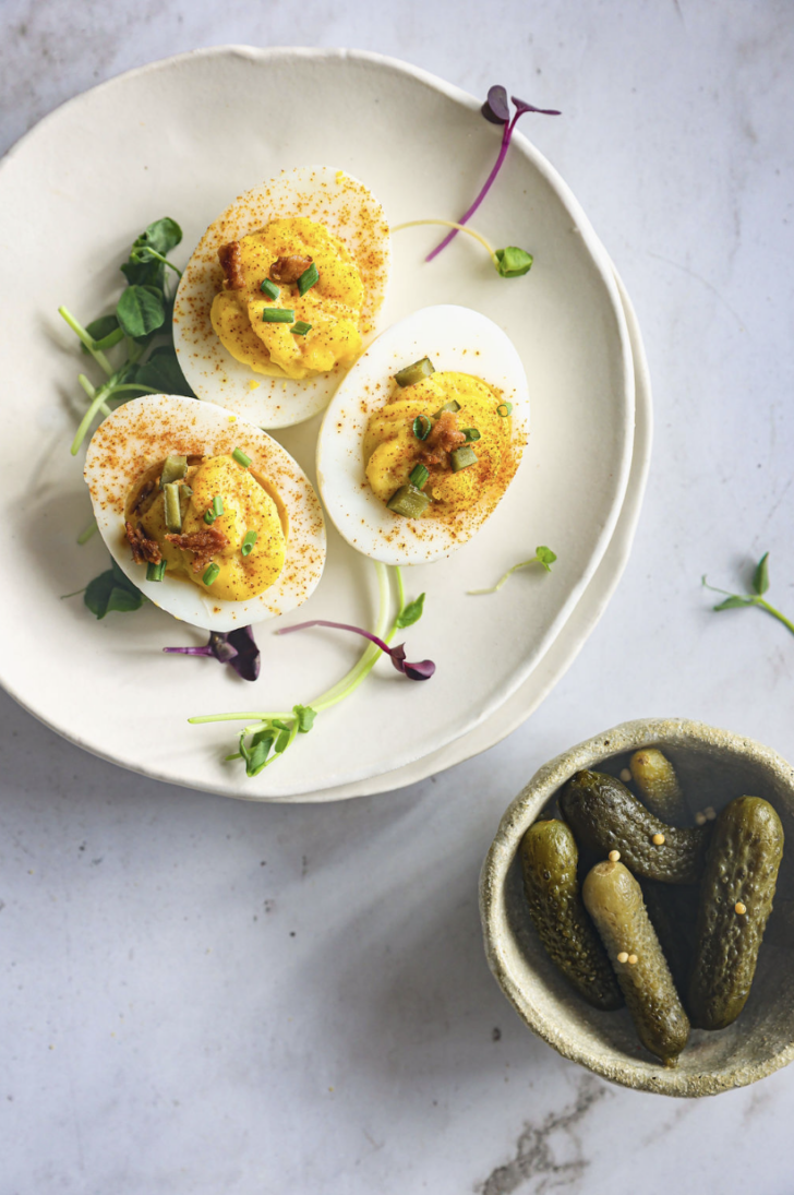 Three deviled eggs garnished with paprika, chives, bacon, and pickles, served on a ceramic plate with microgreens. A bowl of gherkins sits nearby