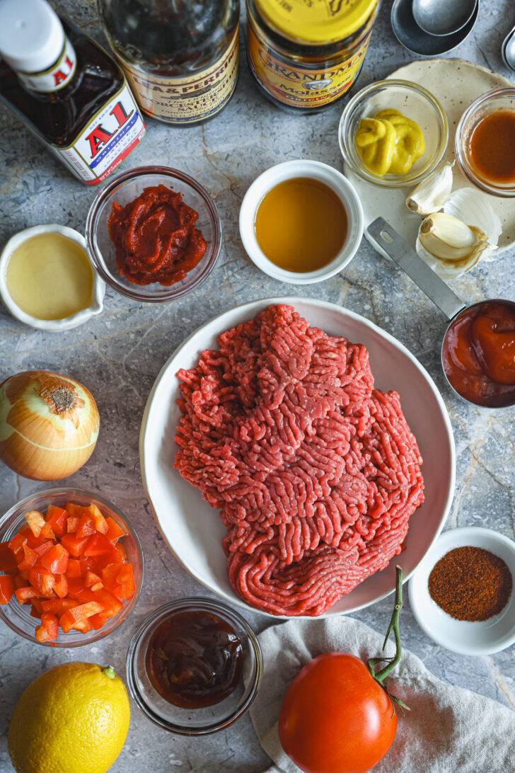 Raw ground beef on a plate surrounded by Sloppy Joe ingredients, including diced bell peppers, onions, tomato paste, mustard, and sauces.