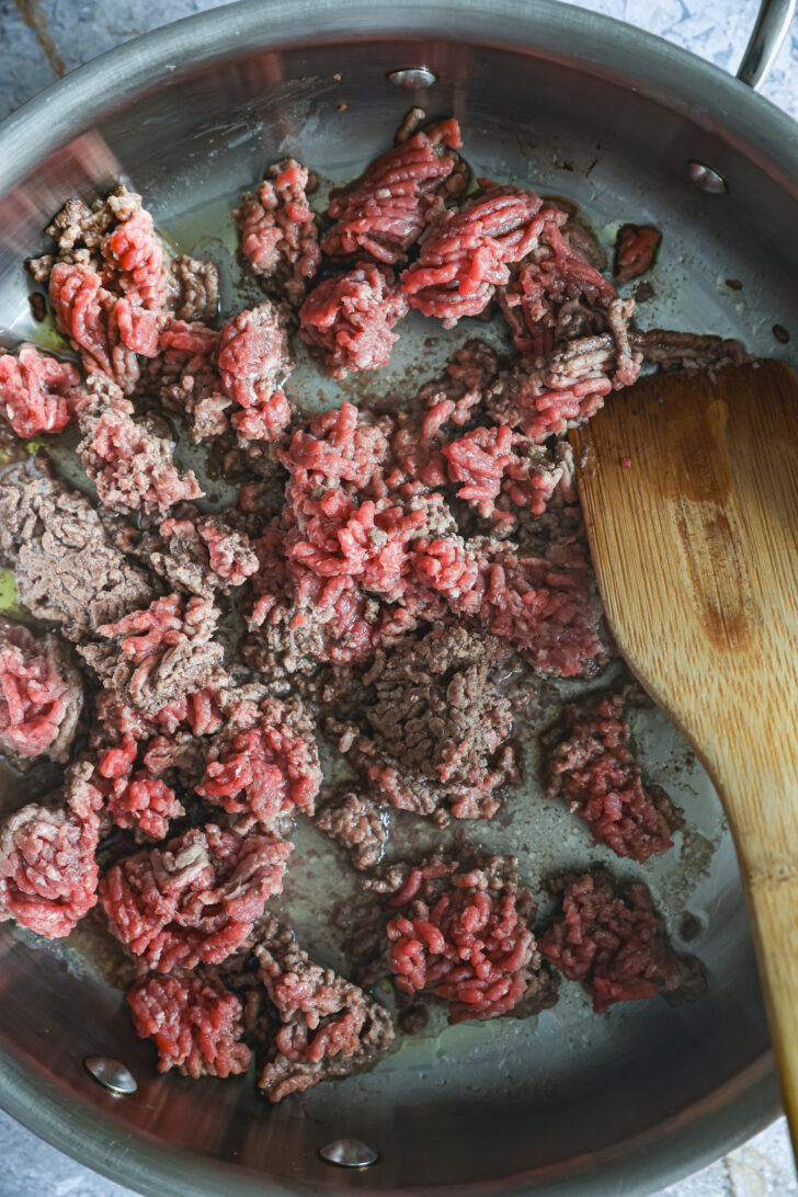 Ground beef browning in a stainless steel skillet with a wooden spatula, partially cooked with some raw pieces remaining.