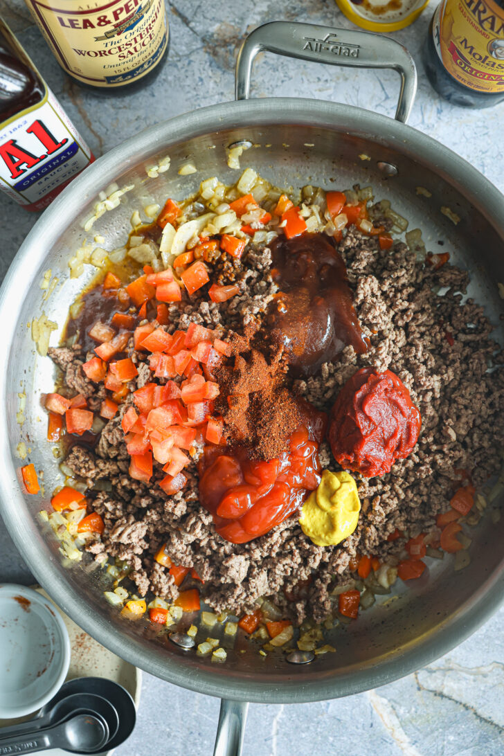 Cooked ground beef in a skillet with diced onions, bell peppers, tomatoes, ketchup, mustard, tomato paste, and seasonings before mixing