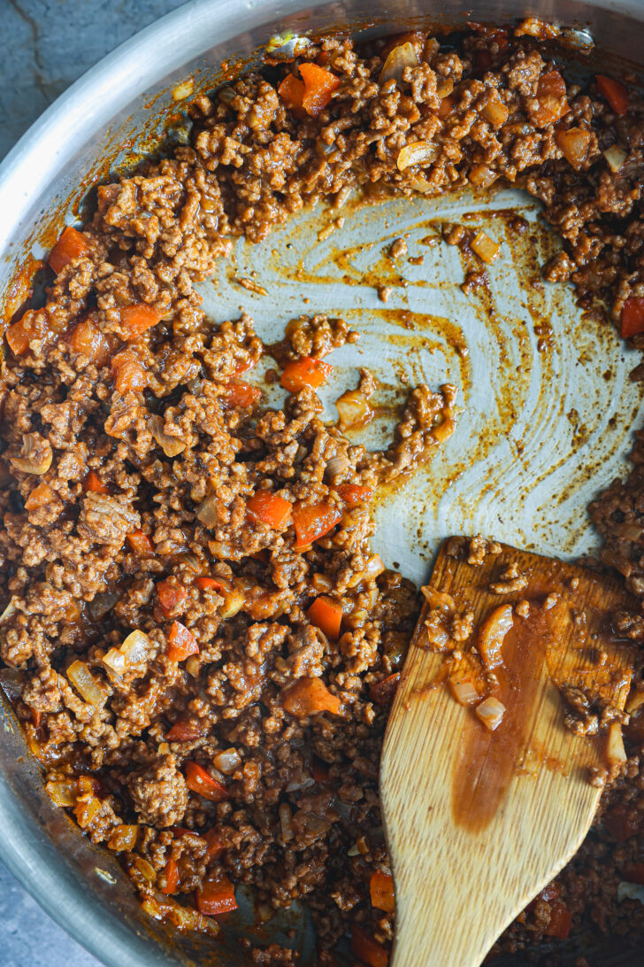 Thick Sloppy Joe filling with ground beef, onions, and peppers simmered in sauce, pushed to the side of a skillet with a wooden spatula.