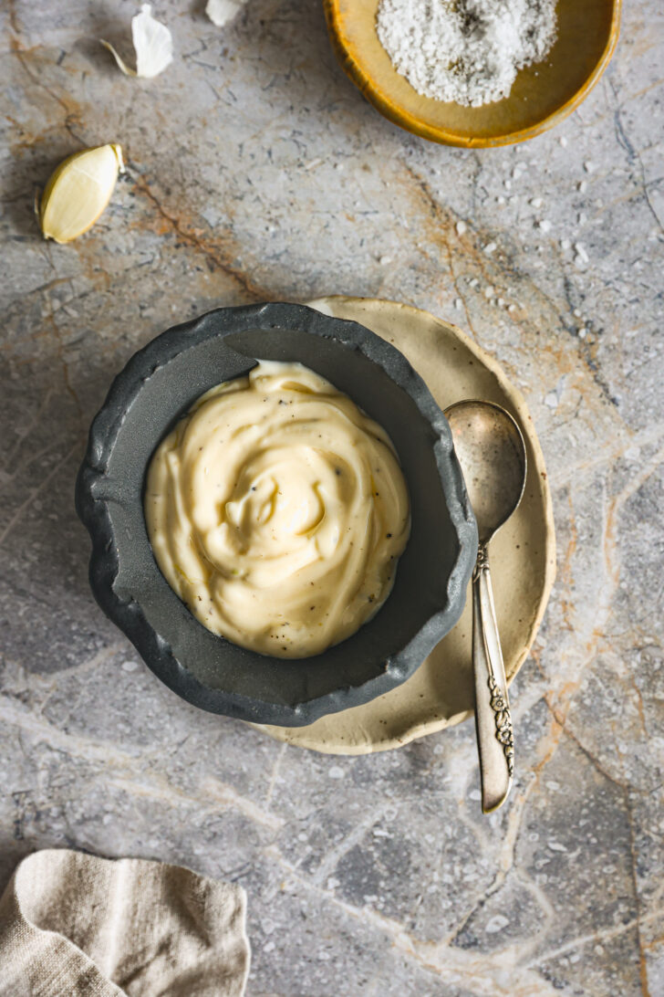 Creamy garlic aioli in a dark stone bowl, placed on a rustic plate with a vintage spoon, surrounded by garlic cloves and coarse salt