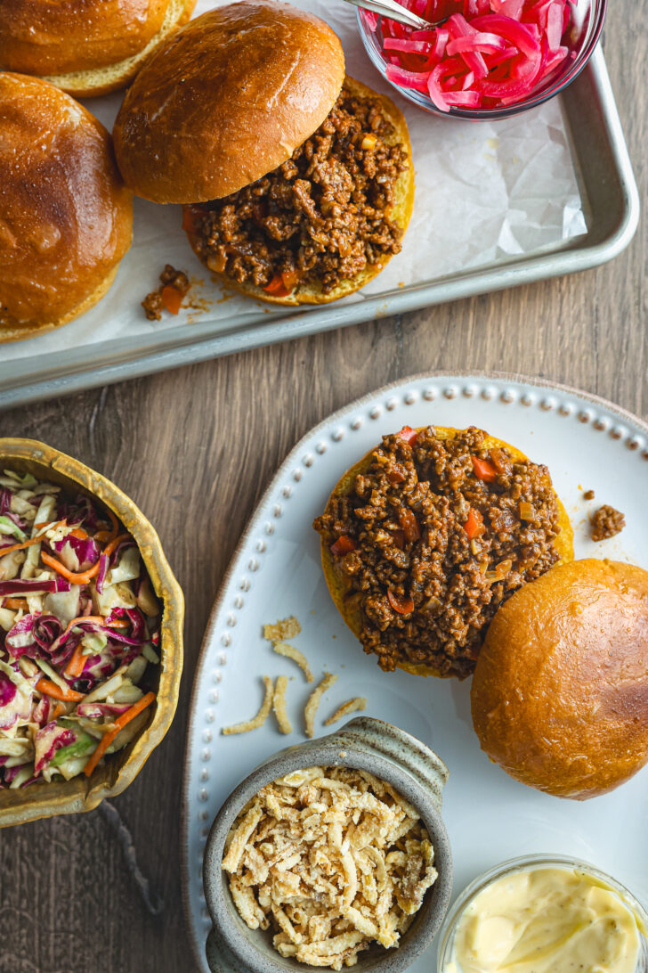 Sloppy Joe sandwiches on brioche buns, served with coleslaw, pickled onions, crispy fried onions, and garlic aioli on a wooden table