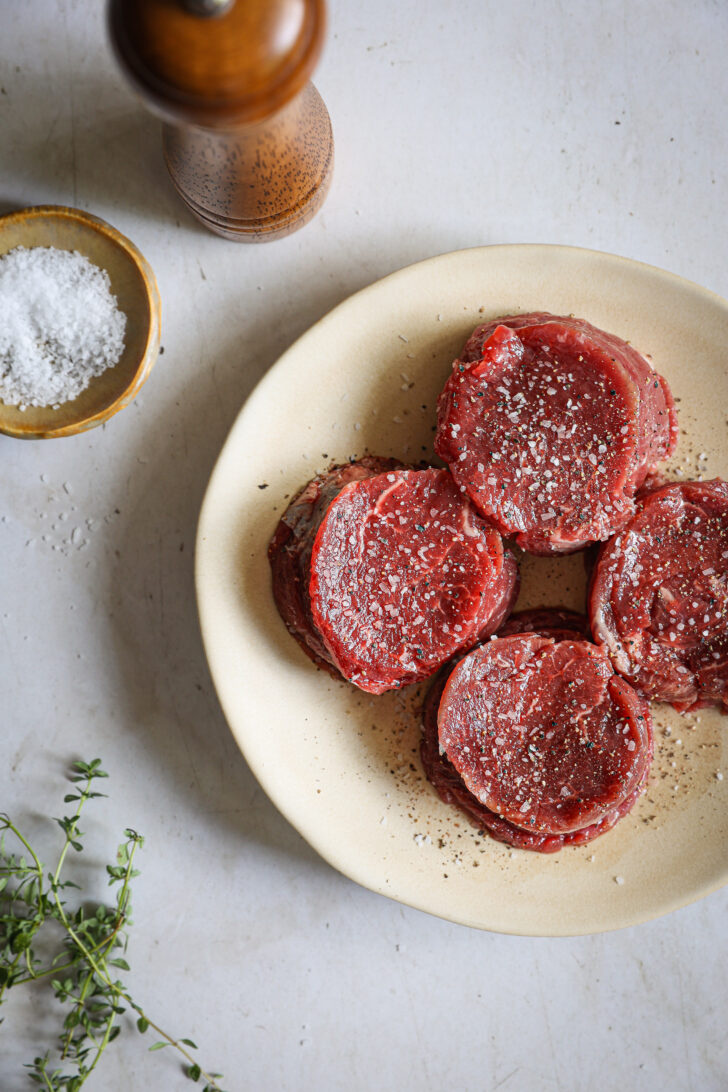 Four raw filet mignon steaks seasoned with kosher salt and cracked pepper on a cream plate, with a pepper mill, salt, and fresh thyme.