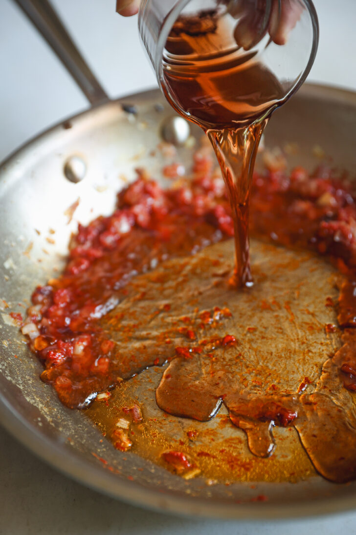 Madeira wine pouring from a glass measuring cup into a stainless skillet with sautéed shallots and tomato paste to deglaze the pan.