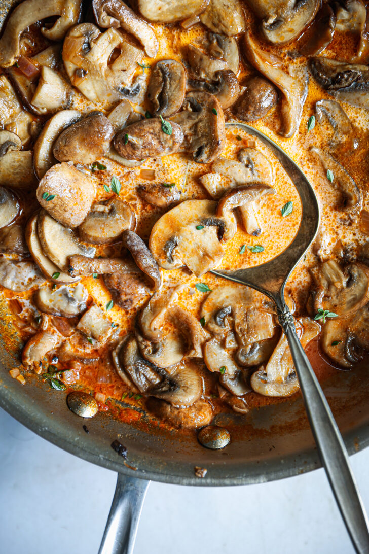 Close-up of finished mushroom Madeira sauce in a skillet, mushrooms simmering in creamy red wine sauce with thyme and a serving spoon.