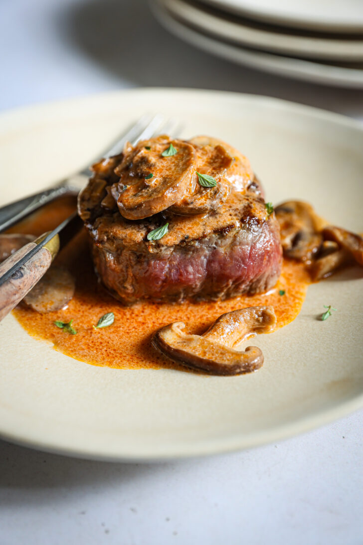 Plated filet mignon topped with mushroom Madeira sauce and fresh thyme, wood-handled steak knife and fork resting alongside.