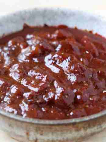 The image shows a bowl of Kansas City-style BBQ sauce. Characterized by its thick consistency and rich, dark color, this sauce is known for a sweet, tangy flavor with a hint of smokiness, typically from ingredients like molasses, tomato paste, vinegar, and spices. The sauce's glossy texture suggests it would be a perfect glaze for meats, offering a sticky, caramelized coating when grilled or baked. The bowl appears to have a rustic finish, which complements the homemade quality of the sauce