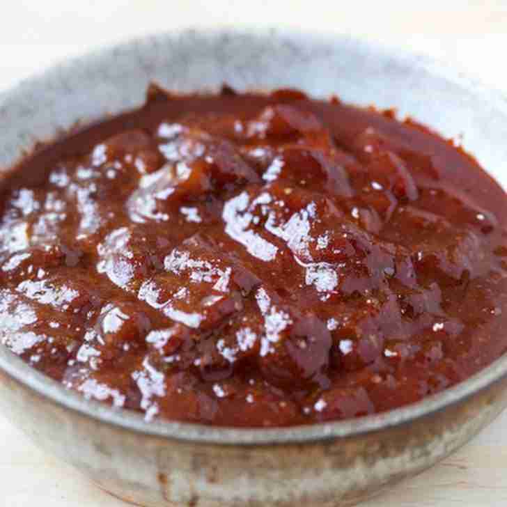 The image shows a bowl of Kansas City-style BBQ sauce. Characterized by its thick consistency and rich, dark color, this sauce is known for a sweet, tangy flavor with a hint of smokiness, typically from ingredients like molasses, tomato paste, vinegar, and spices. The sauce's glossy texture suggests it would be a perfect glaze for meats, offering a sticky, caramelized coating when grilled or baked. The bowl appears to have a rustic finish, which complements the homemade quality of the sauce