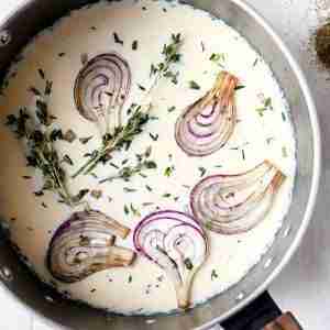 The photo displays a pot filled with infused cream. Slices of red onion are partially submerged in the cream, and fresh thyme sprigs are visible throughout the mixture. The combination of onions and thyme suggests a savory profile for the infusion. The pot has a wooden handle and sits against a light, neutral background which accentuates the contents.