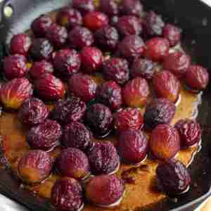 The image depicts a collection of grapes that have been roasted, as evidenced by their shriveled skins and the juices released onto the sheet pan. The grapes are varied in color, ranging from light golden to deep purple, indicating a mix of different grape varieties. The roasting process has caused the grapes to caramelize slightly, and their concentrated sweetness is often used to enhance the flavor profile of both sweet and savory dishes. The sheet pan they rest on has collected some of the juices, which have intensified in color due to the heat exposure.