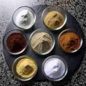 The image captures a top-down view of a lazy susan adorned with seven small glass cups, each containing a different spice. These spices vary in color, suggesting a range of flavors. One cup holds a white powder, likely salt, essential for enhancing the taste of wings. Another contains a bright yellow spice, possibly turmeric or mustard powder, known for its vibrant hue and earthy tones. There's a rich, red spice that could be paprika or chili powder, adding color and heat to the mix. A beige-colored spice, perhaps garlic or onion powder, offers a milder but aromatic quality. There's also a deep brown spice, which might be cumin or clove, known for its warm and intense flavor. The collection of spices on the rotating server suggests a preparation for a flavorful wing seasoning blend, each spice ready to contribute its unique qualities to create a complex and harmonious flavor profile for a batch of wings. The variety of colors from the spices adds a visual appeal to the preparation process, highlighting the culinary artistry involved in seasoning.