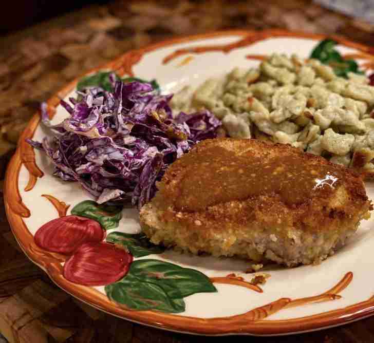 This image presents a beautifully plated Crispy Pork Jägerschnitzel meal. At the center of the plate is a golden-brown, breaded pork cutlet that has been fried to perfection, showing off a crispy exterior. To the right of the schnitzel, there's a serving of Spaetzle, which appears tender and is lightly browned, suggesting it might have been sautéed to add texture and flavor. On the left side of the plate, there's a helping of shredded red cabbage, likely dressed, as indicated by the sheen and slight clinging of sauce to the cabbage, which adds a vibrant contrast in color. The demi-glace, with its rich and glossy look, is elegantly spooned over the schnitzel, giving it a lustrous finish that promises a depth of flavor. The plate itself has a botanical design, contributing to the overall rustic and homey feel of the meal. The food is well-proportioned, suggesting a thoughtful balance between protein, carbs, and vegetables.