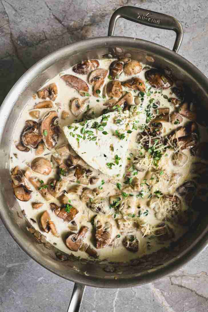 Overhead view of a skillet filled with a creamy mushroom sauce. The sauce includes sliced mushrooms, fresh thyme, and a wedge of blue cheese melting into the mixture. The sauce is rich and creamy, with a sprinkle of grated Parmesan cheese on top, creating a savory base for the gnocchi dish.