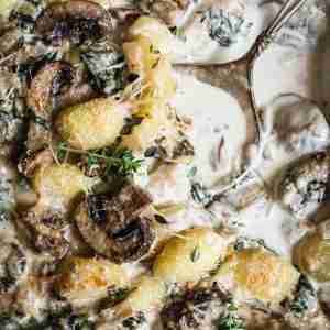 A close-up of creamy baked gnocchi with mushrooms and spinach in a cheese sauce, garnished with fresh thyme sprigs.