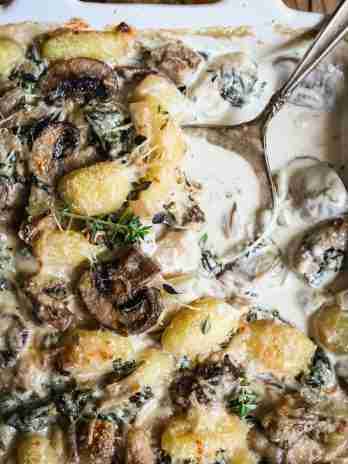 A close-up of creamy baked gnocchi with mushrooms and spinach in a cheese sauce, garnished with fresh thyme sprigs.