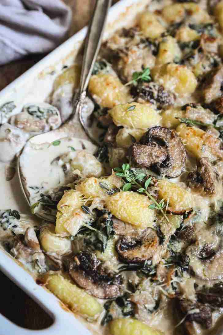 Baked gnocchi with mushrooms and spinach in a creamy sauce, served in a white baking dish. The gnocchi is golden and topped with fresh thyme sprigs. A spoon is nestled in the dish, ready to serve, showing the rich, creamy texture of the sauce surrounding the gnocchi and vegetables.