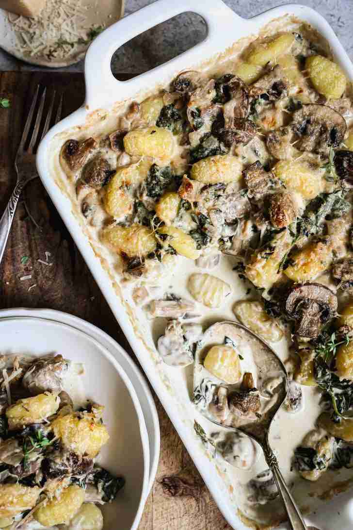 Overhead view of a white baking dish filled with creamy baked gnocchi, mushrooms, and spinach. A serving spoon is placed in the dish, ready to serve. A plate with a portion of the gnocchi is next to the baking dish, with grated Parmesan cheese visible in the background. The dish is garnished with fresh thyme and has a golden, bubbly top.