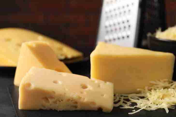 A close-up shot of various blocks of cheese, including Gruyère and Emmental, arranged on a dark surface with a small pile of grated cheese in the foreground. In the background, a metal cheese grater can be seen, emphasizing the preparation process for a dish like cheese fondue. The warm lighting highlights the rich textures and subtle holes in the Emmental, making the cheeses appear fresh and inviting.