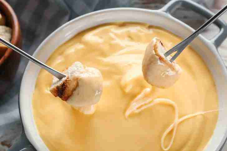 Two metal fondue forks dipping cubes of bread into a pot of smooth, creamy cheese fondue. The melted cheese is a golden yellow, indicating a blend of Gruyère and Emmental, and is perfectly coating the bread. The pot is light-colored, with a rich, velvety texture to the cheese, making it look warm and inviting.