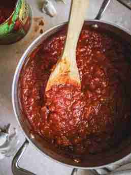 Overhead view of a pot filled with rich, thick tomato sauce being stirred with a wooden spoon. The sauce is made from crushed tomatoes and has a deep red color, with visible chunks of onion and garlic. An open can of crushed tomatoes and garlic cloves are seen in the background.