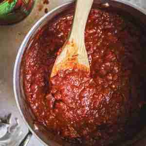 Overhead view of a pot filled with rich, thick tomato sauce being stirred with a wooden spoon. The sauce is made from crushed tomatoes and has a deep red color, with visible chunks of onion and garlic. An open can of crushed tomatoes and garlic cloves are seen in the background.