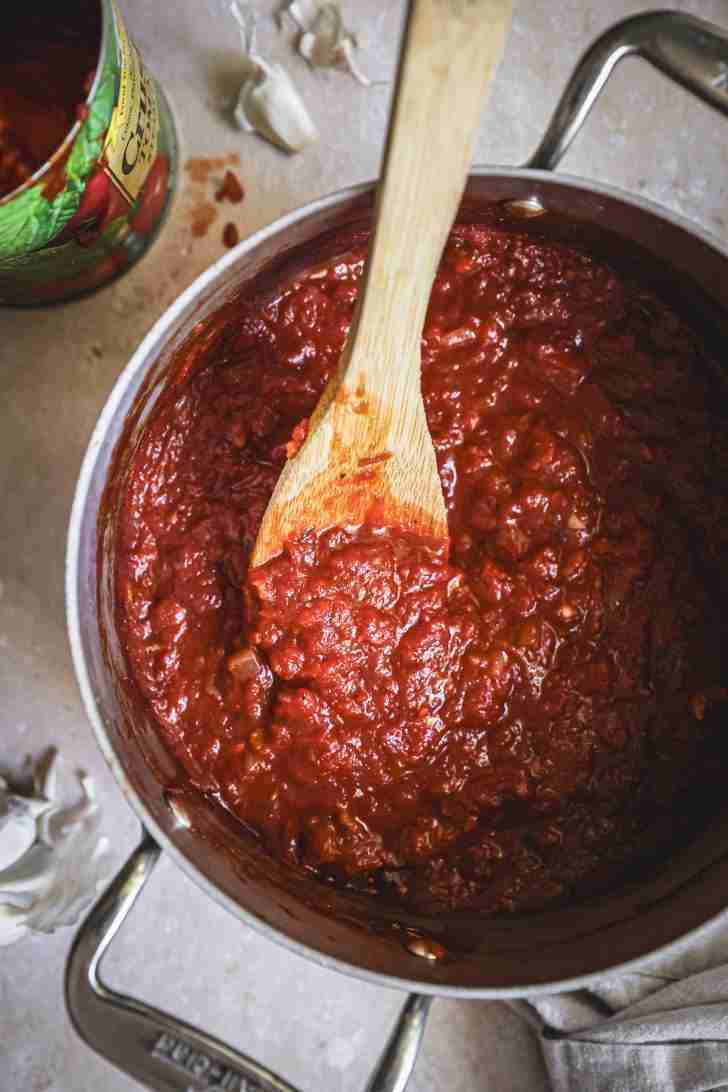 Overhead view of a pot filled with rich, thick tomato sauce being stirred with a wooden spoon. The sauce is made from crushed tomatoes and has a deep red color, with visible chunks of onion and garlic. An open can of crushed tomatoes and garlic cloves are seen in the background.