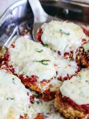 Close-up of freshly baked Chicken Parmesan in a baking dish. The chicken breasts are coated with a crispy breadcrumb crust, topped with marinara sauce, melted mozzarella, and Parmesan cheese. Fresh basil is sprinkled on top, and a spatula is seen lifting one piece, showing the gooey, melted cheese.