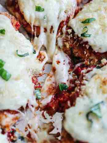 Close-up of Chicken Parmesan in a baking dish, showing melted mozzarella cheese being pulled apart with a stringy, gooey texture. The chicken is topped with marinara sauce and garnished with fresh basil, with a crispy breadcrumb coating peeking through.