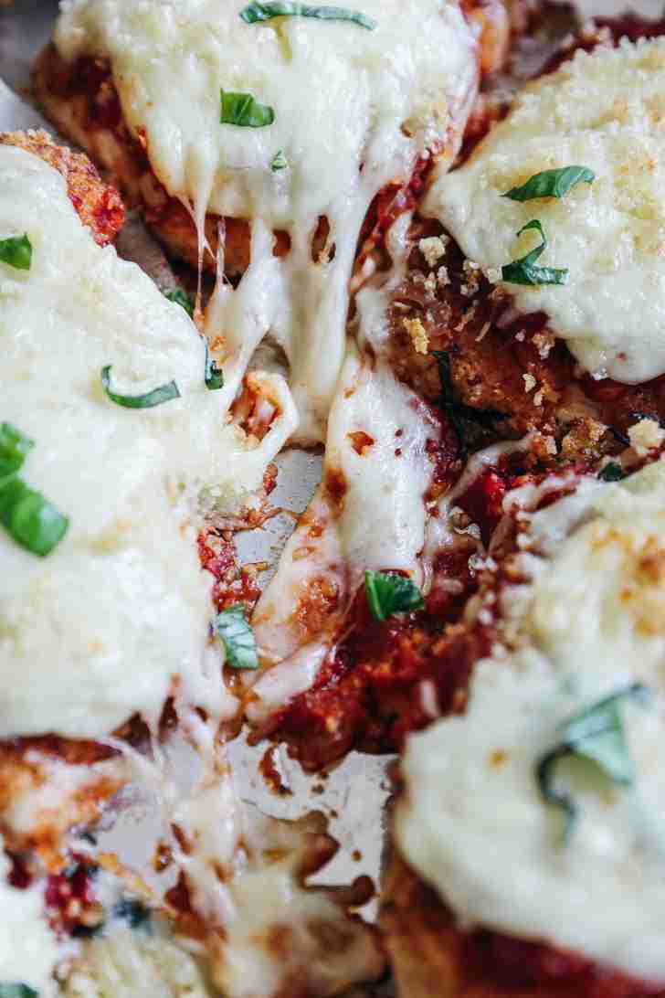 This is a mouth-watering image of Chicken Parmesan, freshly prepared and ready to be served. Six golden-brown, breaded chicken fillets are neatly arranged on a wooden cutting board. Each fillet is topped with a rich and hearty red marinara sauce, oozing with flavor. Over the sauce, there's a generous portion of melted mozzarella cheese that has been broiled to perfection, creating a bubbly and slightly golden finish. Sprinkled on top of the cheese, you can see a dusting of finely chopped herbs, likely basil or parsley, adding a pop of color and fresh flavor to the dish. The presentation is rustic yet inviting, suggesting that the meal is homemade with care and attention to both taste and presentation.