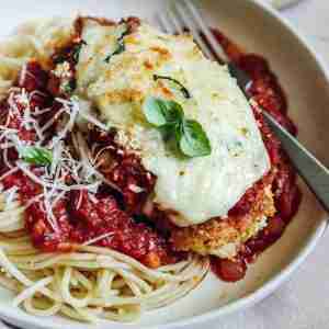 A plated serving of Chicken Parmesan on a bed of spaghetti topped with marinara sauce. The chicken is covered in melted mozzarella and garnished with fresh basil leaves. Grated Parmesan cheese is sprinkled over the spaghetti, and a fork rests beside the dish, ready to enjoy.